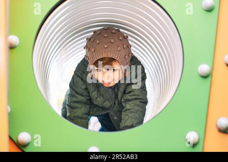 Ragazza carina del bambino che ha divertimento sul parco giochi, che striscia attraverso il tubo, tempo freddo Foto Stock