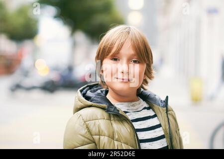 Ritratto all'aperto di un bel ragazzo di 10 anni, che indossa una giacca calda, guarda dritto la macchina fotografica, che si posa per strada Foto Stock