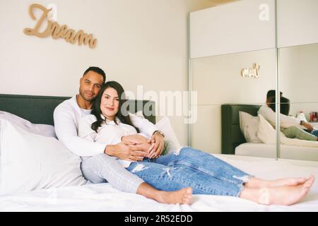 Ritratto interno di felice giovane famiglia, donna incinta con il suo marito amorevole rilassarsi a letto Foto Stock