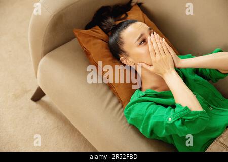 Giovane donna che piange sdraiata su un divano a casa copra il viso con le mani. Foto Stock