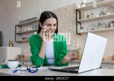 Una giovane donna si siede a casa in cucina di fronte a un portatile e grida, le asciuga le lacrime con un tovagliolo, punta la mano sullo schermo, comunica durante una videochiamata, legge le notizie, ha ricevuto cattive notizie. Foto Stock