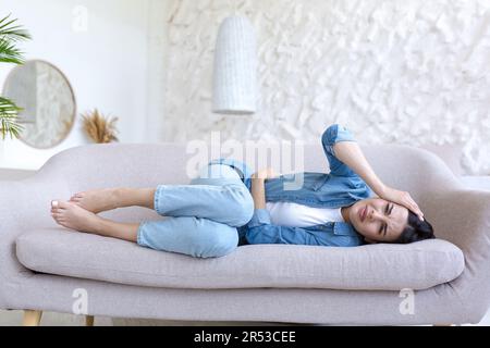Una giovane donna si trova a casa, in un albergo, su un divano e tiene lo stomaco e la testa con le mani. Soffre di dolore, tensione, pressione. Ha bisogno di assistenza medica. Foto Stock