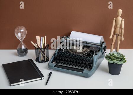 Macchina da scrivere vintage con manichino in legno, portamatite e clessidra su tavolo bianco vicino alla parete marrone Foto Stock