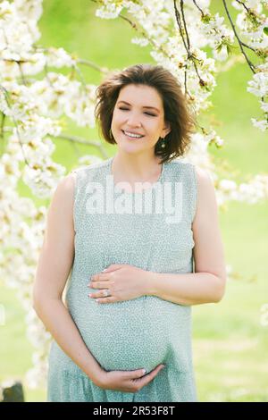 Ritratto di maternità all'aperto di felice giovane donna incinta in piedi accanto all'albero fiorente, primavera Foto Stock