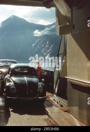 Geirangerfjord, Norvegia. Circa. 1960 – Una giovane donna in piedi accanto a un Beetle Volkswagen sul ponte di un traghetto sul Geirangerfjord. Foto Stock