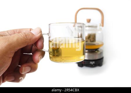 Foto ravvicinata di una mano che tiene un piccolo teacup di vetro con teacettle sfocato su sfondo bianco. Foto Stock