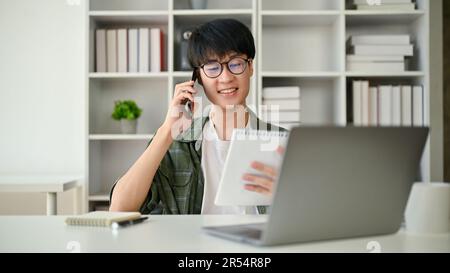 Un giovane assistente asiatico in occhiali ha guardato i dettagli su un blocco note mentre parlava al telefono con un cliente aziendale alla sua scrivania. Foto Stock