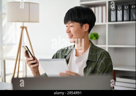 Un giovane uomo d'affari asiatico felice o assistente di vendita maschile sta digitando al telefono, inviando messaggi, o chiamando qualcuno mentre si siede alla sua scrivania. Foto Stock
