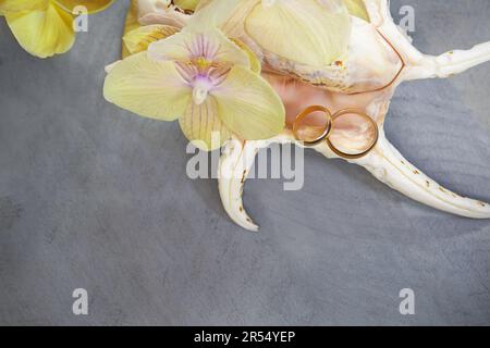 Due anelli da sposa dorati in grande conchiglia con orchidea gialla su fondo grigio in legno. Cerimonia nuziale in paesi caldi esotici. Spazio di copia Foto Stock