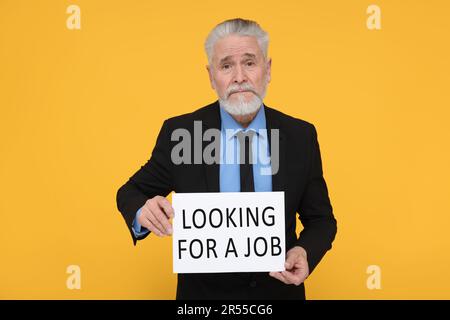 Uomo anziano disoccupato con segno di cartone con frase cercando Un lavoro su sfondo giallo Foto Stock