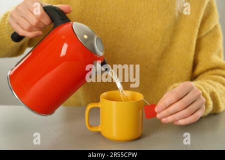 Donna che versa acqua calda in tazza con sacchetto di tè al tavolo grigio, primo piano Foto Stock