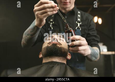 Parrucchiere con olio di barba presso il cliente in barbiere, primo piano. Servizio di rasatura professionale Foto Stock