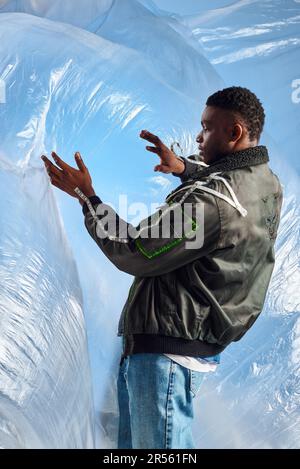 Vista laterale dell'elegante uomo afroamericano in giacca con righe a LED e jeans strappati che toccano il cellophane lucido su sfondo blu, outfit urbano e. Foto Stock