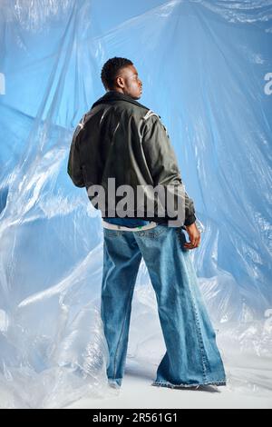 Vista laterale di un uomo afroamericano sicuro ed elegante in giacca outwear con righe a LED e jeans in piedi vicino cellophane su sfondo blu, urbano ou Foto Stock
