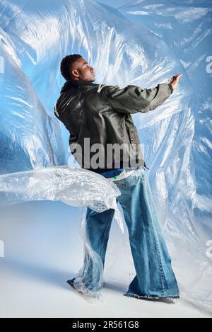 Vista laterale di moda giovane afroamericana modello maschile in giacca outwear con righe a LED e jeans in posa su lucido cellophane su sfondo blu, ur Foto Stock