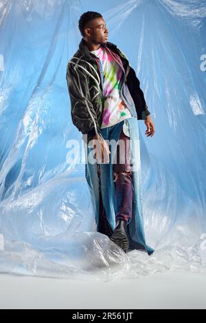 Uomo alla moda afroamericano in giacca con righe a LED e jeans strappati che cammina su cellophane lucido su sfondo blu, abito urbano e mod Foto Stock