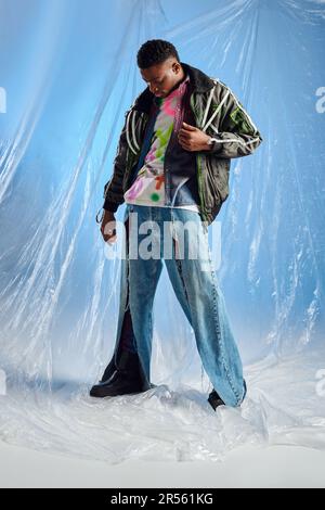 Bel giovane uomo afroamericano in giacca outwear con righe a LED guardando jeans strappati alla moda in piedi su cellophane lucido su sfondo blu, ur Foto Stock