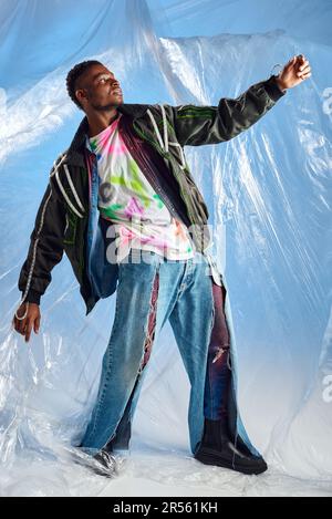 Uomo alla moda afroamericano in giacca outwear con righe a LED e jeans strappati in posa su cellophane lucido su sfondo blu, outfit urbano e modalità Foto Stock