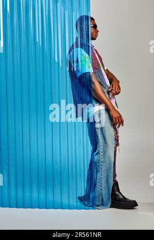 Vista laterale di un elegante modello afroamericano maschio in jeans strappati, giubbotto denim e occhiali da sole in piedi dietro un foglio di policarbonato blu su sfondo grigio, D Foto Stock