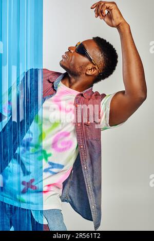 Vista laterale del giovane uomo alla moda afroamericano in occhiali da sole, gilet in denim colorato e t-shirt posate e in piedi dietro un foglio di policarbonato blu sul grigio Foto Stock