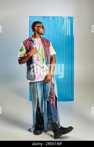 Vista laterale di un elegante uomo afroamericano in occhiali da sole, gilet denim colorato e jeans strappati in posa grigia con foglio di policarbonato blu sullo sfondo, Foto Stock