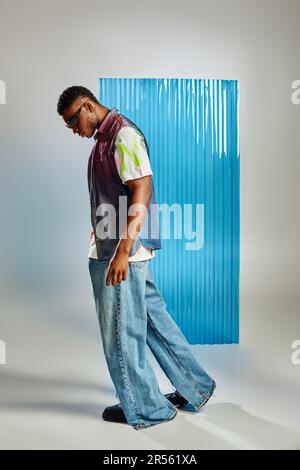 Vista laterale di un elegante modello afroamericano in occhiali da sole, giubbotto in denim e jeans che camminano in grigio con foglio di policarbonato blu sullo sfondo, Sostenab Foto Stock