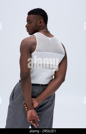 Vista laterale del giovane atletico afro-americano in canotta top e pantaloni guardando lontano e posando con fiducia in elegante e alla moda vestito isolato su gr Foto Stock