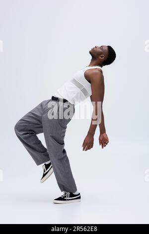 Vista laterale di muscoloso modello afroamericano in canotta top e pantaloni che posano con fiducia in elegante e alla moda vestito su sfondo grigio, buon aspetto Foto Stock