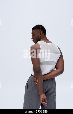 Vista laterale del giovane uomo afro-americano muscoloso in pantaloni e t-shirt senza maniche che si posano con fiducia in un abito elegante e alla moda isolato sul grigio, goo Foto Stock