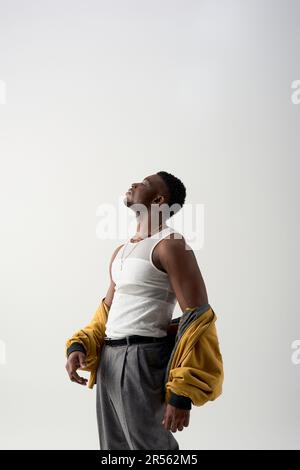 Vista laterale di un giovane afro-americano forte e alla moda in canotta e giacca bomber in piedi isolato su grigio, sparo contemporaneo con stylis Foto Stock