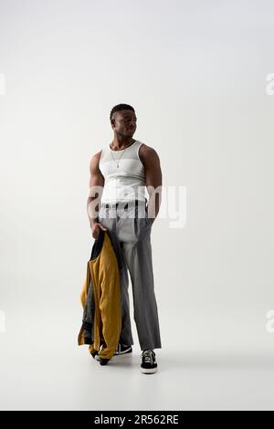 Tutta la lunghezza del giovane uomo afroamericano alla moda in t-shirt senza maniche e pantaloni che tengono la giacca bomber su sfondo grigio, sparatutto contemporaneo Foto Stock