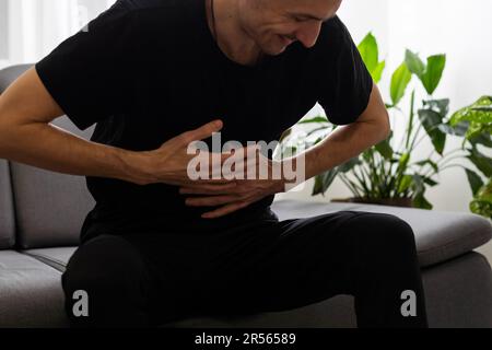 L'uomo è seduto su un divano grigio e tiene il ventre. Medicina e concetto di salute, problemi di stomaco. L'uomo soffre di mal di stomaco, gastrico Foto Stock