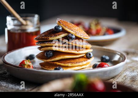 Pancake americani con mirtilli, fragole e ciliegie con salsa di miele. Posto su un piatto rustico di design, su un tavolo rustico in legno. Foto Stock