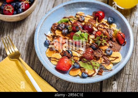 Piccoli pancake americani mescolati con frutta di stagione come fragole, mirtilli e ciliegie. Ricoperto di cioccolato fuso, posto su un legno rustico Foto Stock