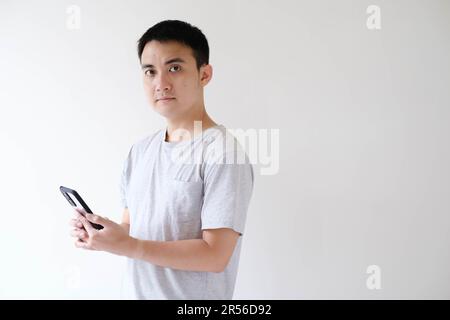 Un giovane asiatico che indossa una t-shirt grigia tiene uno smartphone e guarda la fotocamera. Sfondo bianco isolato. Foto Stock