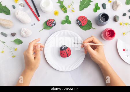 Disegnare una coccinella su una pietra con vernici acriliche. Gli hobby domestici sono autentici. Opere d'arte su pietre. Mani di donna. Foto Stock