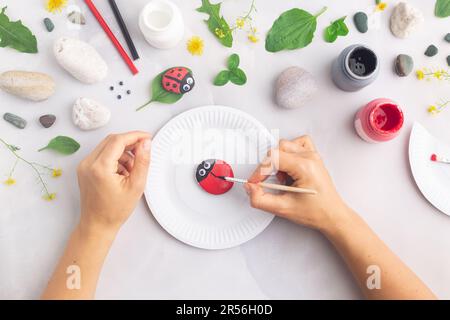 Disegnare una coccinella su una pietra con vernici acriliche. Gli hobby domestici sono autentici. Opere d'arte su pietre. Mani di donna. Foto Stock