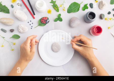 Disegnare una coccinella su una pietra con vernici acriliche. Gli hobby domestici sono autentici. Opere d'arte su pietre. Mani di donna. Foto Stock