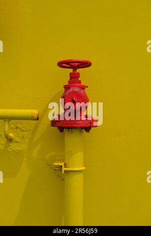 Una valvola dell'idrante verniciata di rosso con una ruota per chiudere e aprire il flusso d'acqua. Valvola dell'idrante antincendio verniciata di rosso su una parete gialla. Foto Stock