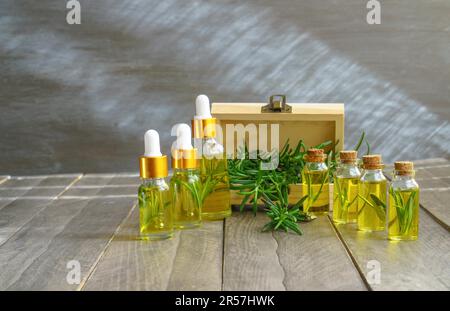 Bottiglie in fila con contagocce di olio essenziale di rosmarino con una scatola di legno e rami di rosmarino fresco, la luce del sole arriva attraverso la finestra Foto Stock