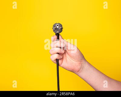 Una mano contiene una spina a cui è collegato un cavo nero. Senza volto, sfondo giallo. Foto Stock