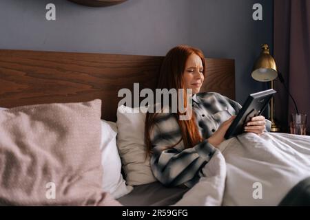 Vista laterale di una giovane donna afflicata sdraiata sul letto tenendo la foto in cornice e piangendo gli occhi chiusi, toccando l'immagine con amore a casa. Foto Stock