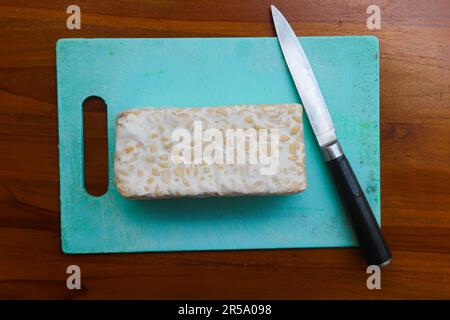 Tempeh crudo fresco, su un tagliere e coltello per tritare. Concetto di preparazione del cibo Foto Stock