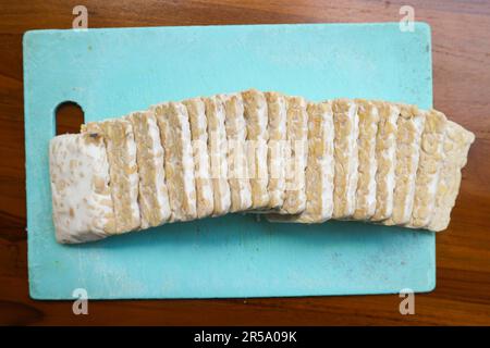 Tempeh crudo fresco a fette, su un tagliere di plastica su uno sfondo di legno. Concetto di preparazione del cibo Foto Stock