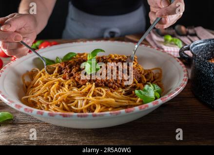 Donna con grembiule sta mescolando spaghetti con ragu alla bolognese in un grande recipiente per pasta su un tavolo di legno Foto Stock