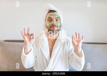 L'uomo divertente gode di trattamenti di bellezza e relax di lusso presso la spa di casa. Un uomo con un asciugamano sulla testa siede su un divano in un accappatoio bianco e con un Foto Stock