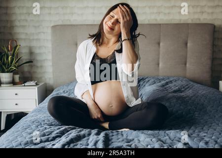 Ragazza incinta seduta sul letto con gli occhi chiusi Foto Stock