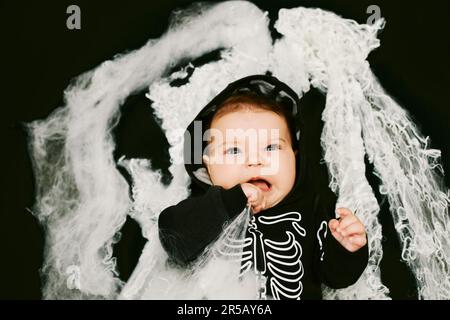 Ritratto di Halloween di adorabile bambino indossando costume scheletro, posando su sfondo nero ricoperto di ragnatela Foto Stock