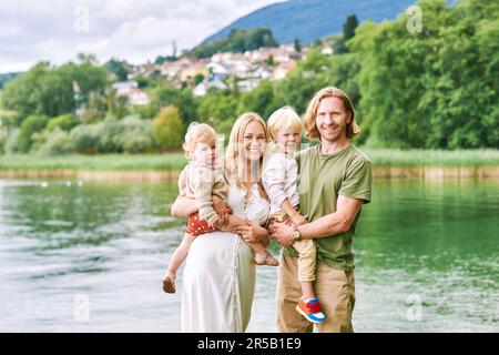 Ritratto all'aperto di bella famiglia, giovane coppia con bambino preschooler e bambina in posa accanto al lago o fiume Foto Stock