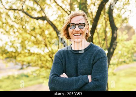Ritratto all'aperto di bell'uomo di 35 - 40 anni con capelli rossi, in posa in verde parco soleggiato, con indosso un pullover blu e occhiali da sole Foto Stock